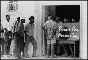 A group of African-American men stand outside a building protesting the segregation of swimming pools in Cairo, Illinois. Inside the entrance, a group of white men stand behind a sign that reads "Private Pool: Members Only."