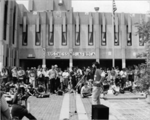 David Roediger is pictured speaking before a large crowd assembled outside a concrete building. In the background, a sign plastered to the building reads "NO BUSINESS IN S. AFRICA."