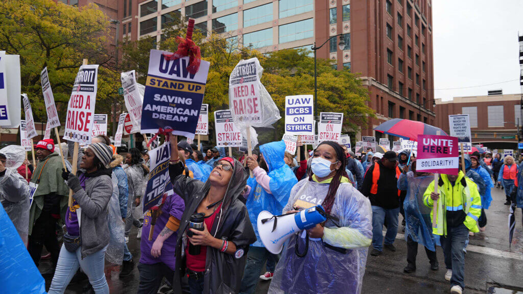A crowd of protesters marches down a city street. In the center, a woman carries a megaphone. Many of the protesters carry picket signs with the UAW logo.